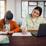 A woman and child writing at a desk beside a laptop.