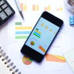 A phone displaying charts on top of printed reports beside a calculator.