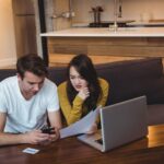 Two people reviewing papers with a laptop and phone in a living room.